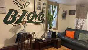 A living room with a wall mural reading "BLOC," a coffee table with books and a vase, a sofa with orange pillows, and a window with plants. - B-Love's Guest House (Oakland)