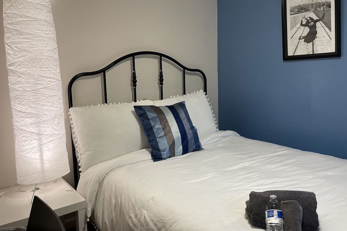 Un lit bien fait, avec des oreillers à rayures blanches et bleues, une lampe blanche et un tableau encadré accroché au mur.