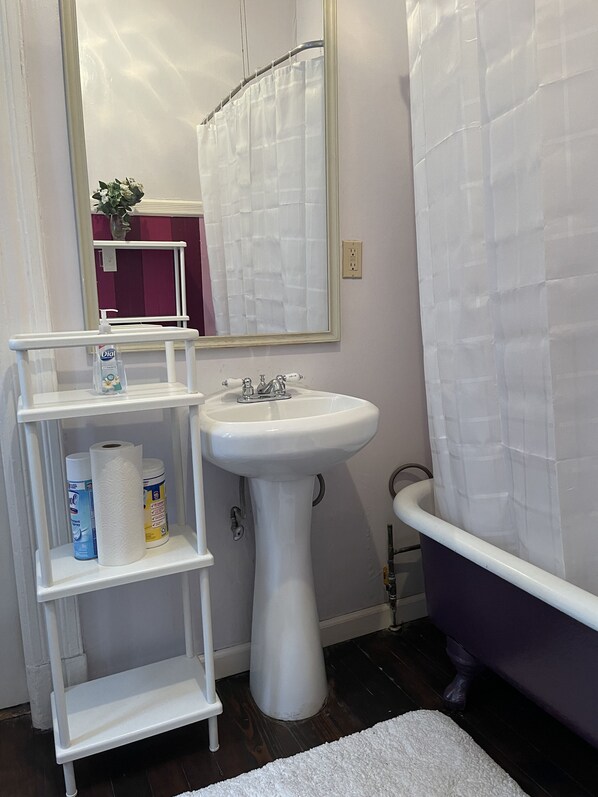 Un baño con lavamanos pedestal, un estante blanco con artículos de aseo, un espejo y una bañera.