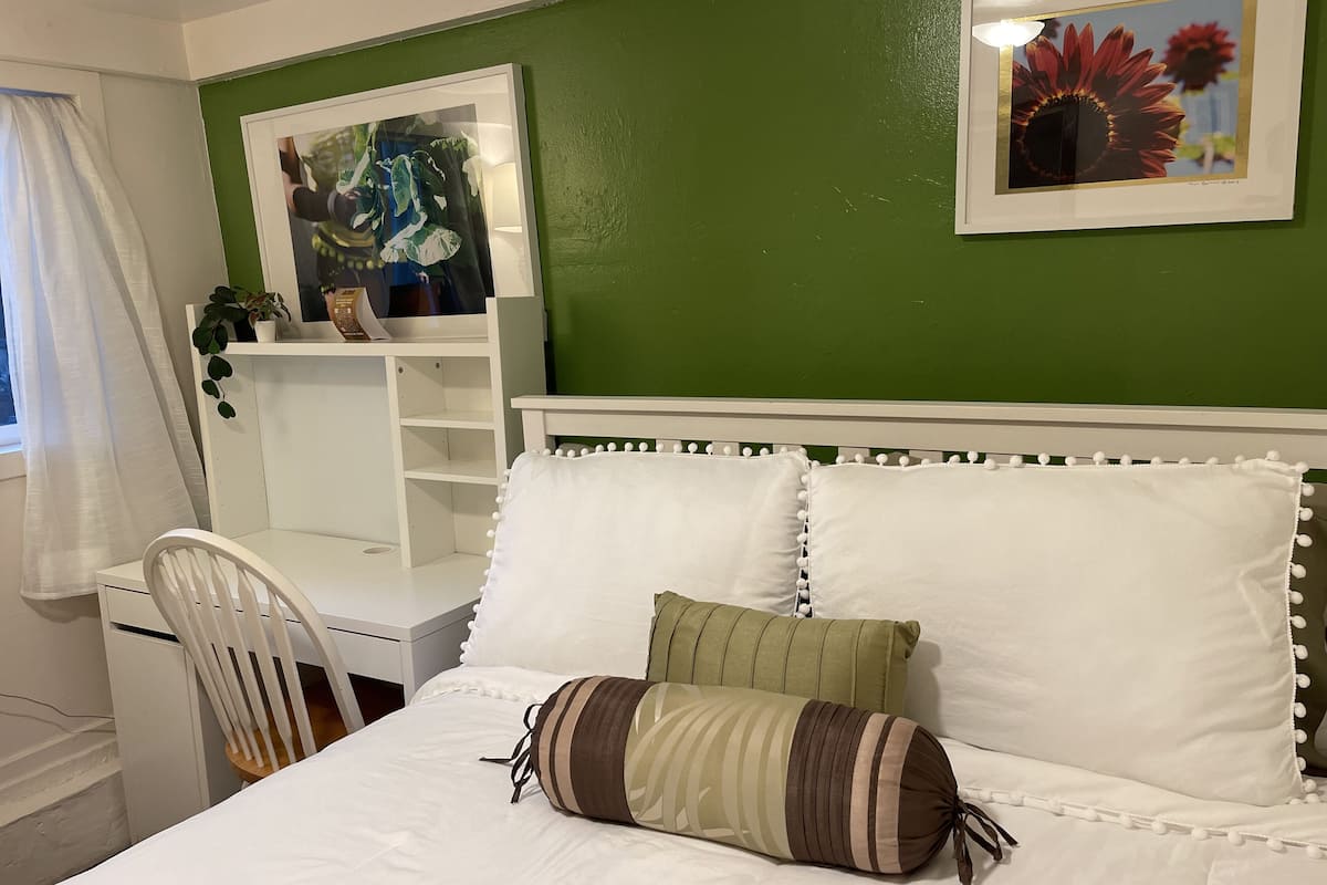 Une chambre à coucher avec un lit, une tête de lit blanche et des oreillers, un bureau avec une chaise, un mur vert et deux tableaux encadrés.