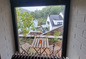 Blick aus einem Fenster, draußen ein Holztisch und Stühle, ein weißes Gebäude und ein Baum mit grünen Blättern.
