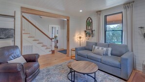 A spacious living room with a grey sofa, a brown leather armchair, and a wooden coffee table. A staircase leads to an upper floor, and a large window provides natural light.