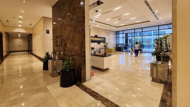 Um hall espaçoso com balcão de recepção em mármore, plantas em vasos e uma grande janela com vista para o exterior.