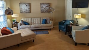 Een woonkamer met een beige bank, een blauwe fauteuil, een vloerlamp en twee aan de muur bevestigde schilderijen.