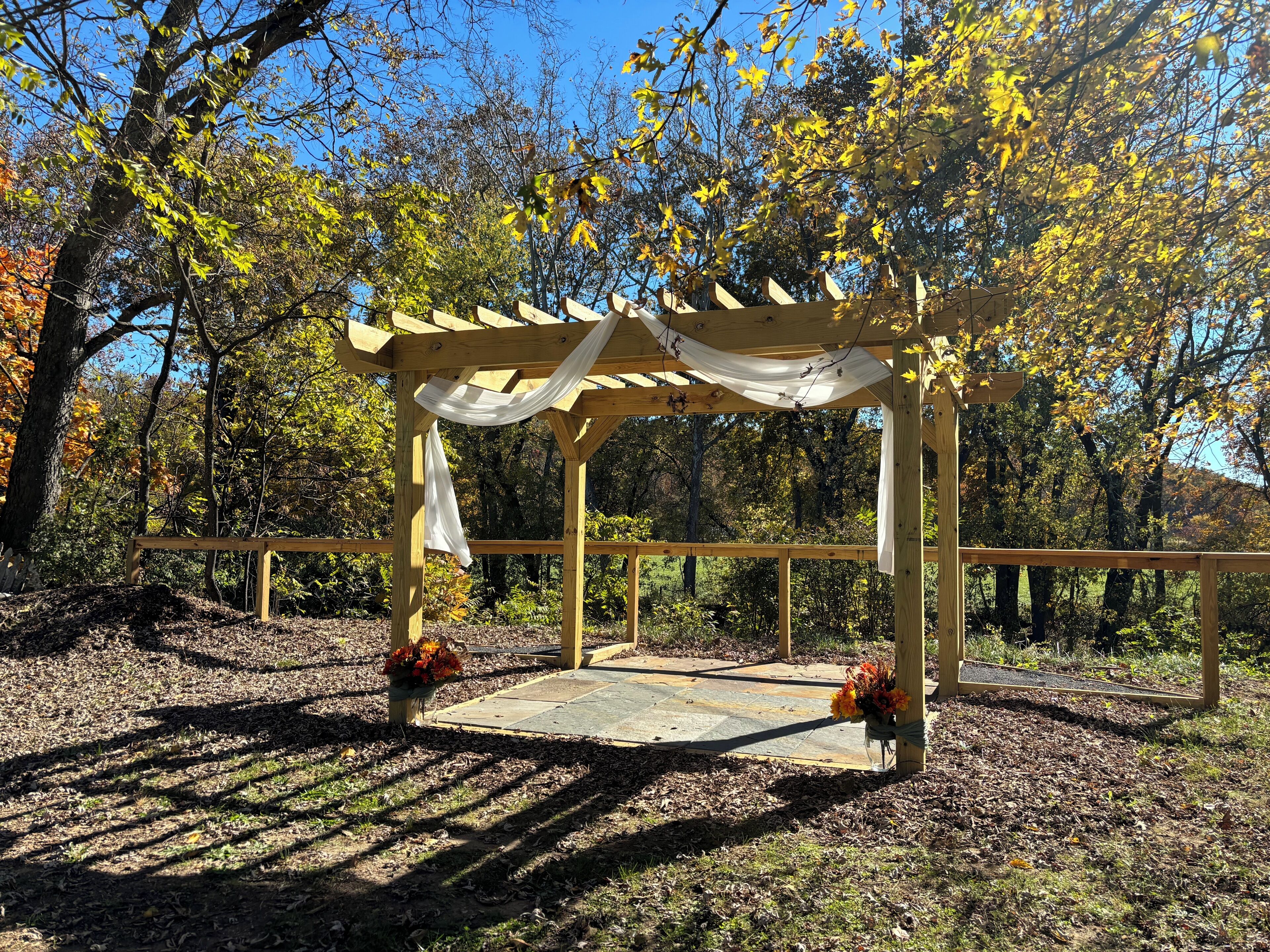 A wooden gazebo with a white draped canopy, surrounded by trees with autumn foliage.