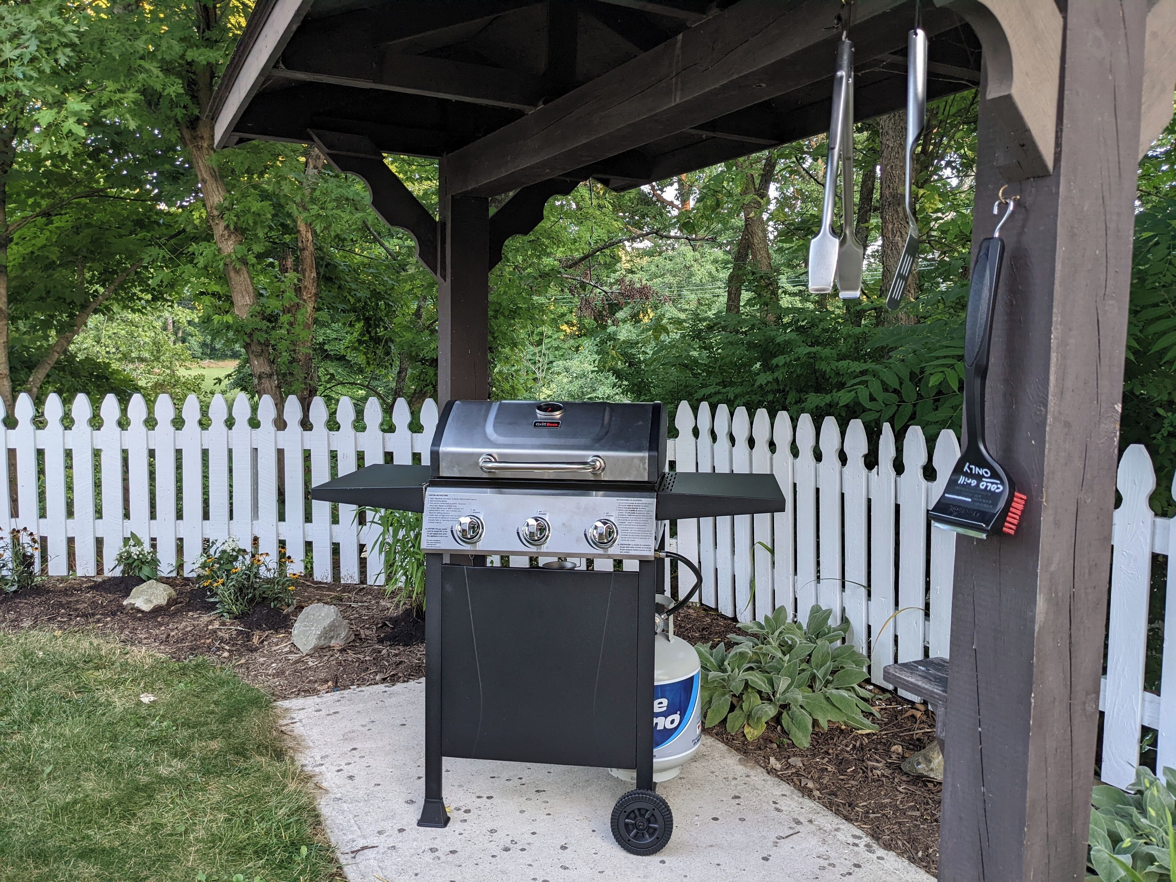 Een barbecue onder een houten prieel, met een witte schutting op de achtergrond.