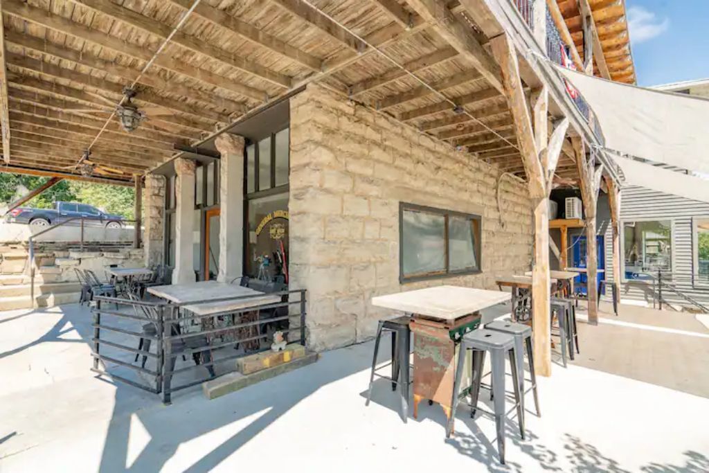 A rustic building with a stone facade, a covered patio area with metal tables and chairs, and a wooden ceiling structure.