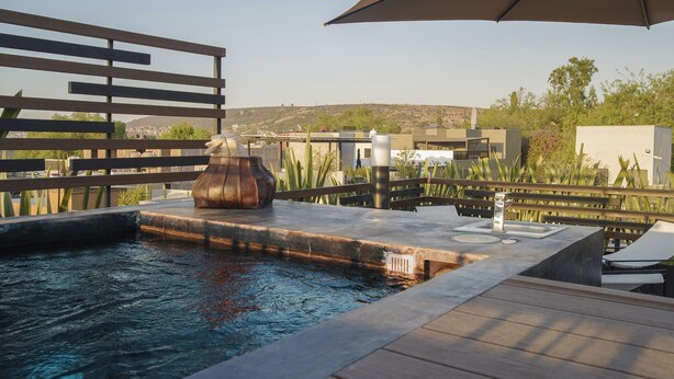 Un área de piscina con terraza de madera, una escultura de metal y una pérgola que ofrece sombra.