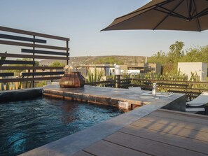 Un área de piscina con terraza de madera, una escultura de metal y una pérgola que ofrece sombra.