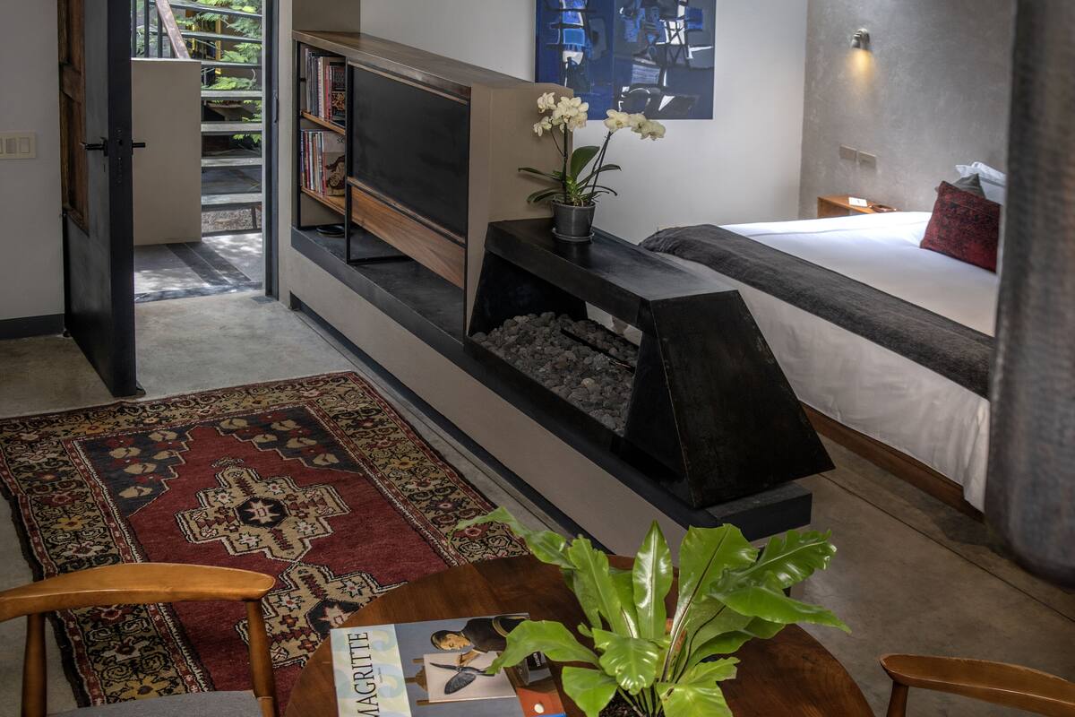 A modern living room with a sofa, a coffee table with a book, a fireplace, a bed, and a bookshelf.