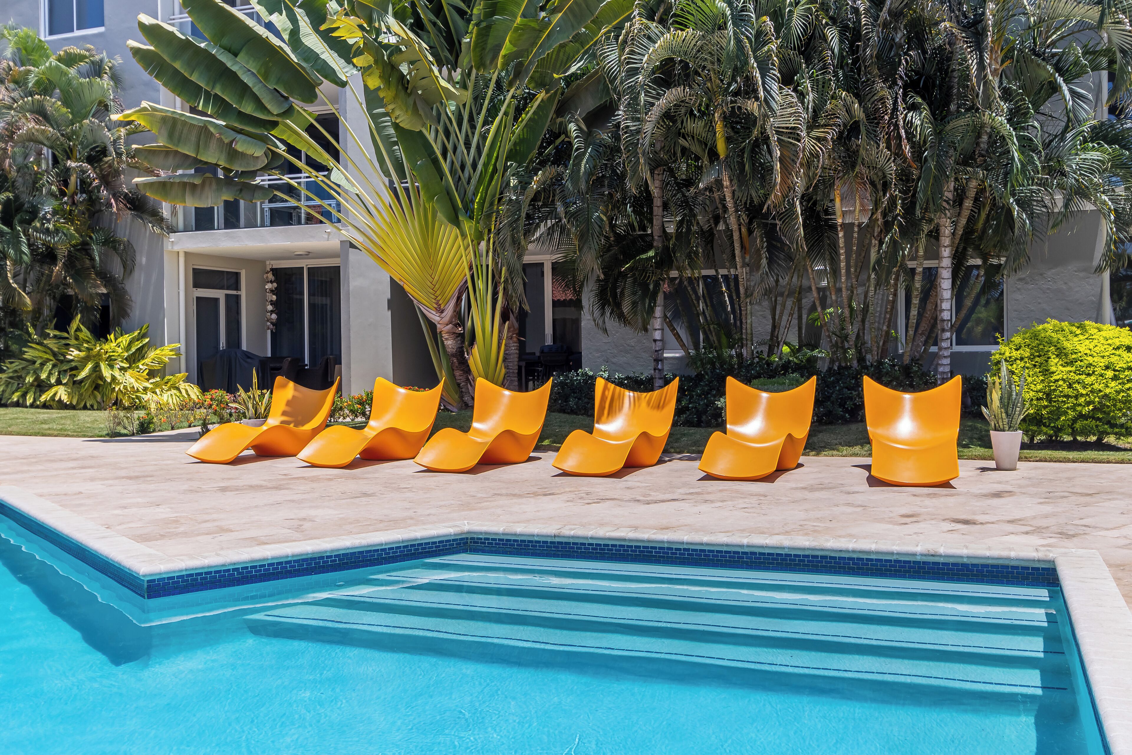 A pool area with a clear blue pool, surrounded by orange lounge chairs and palm trees.