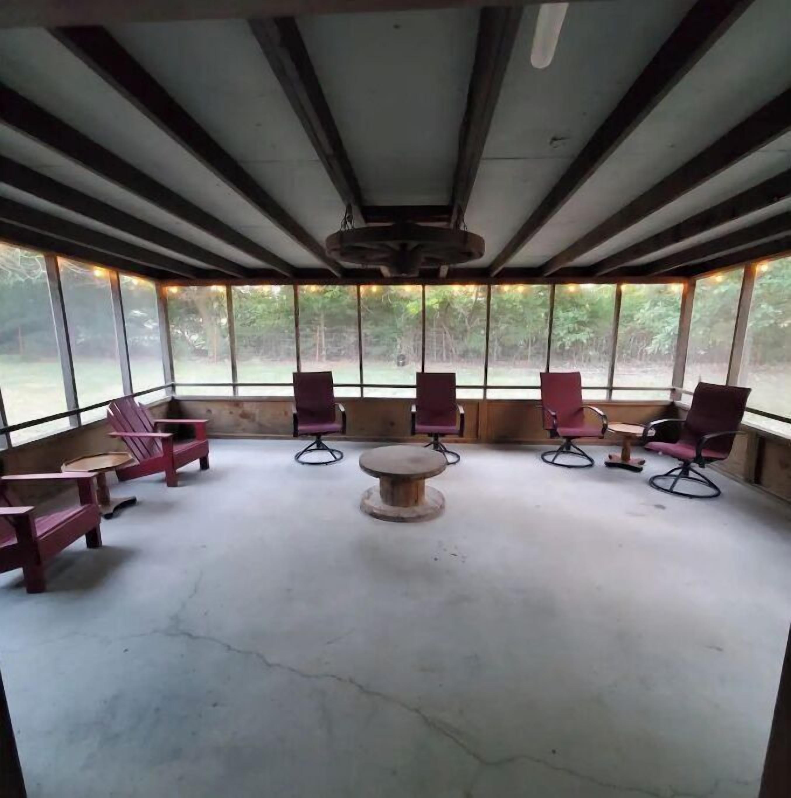A covered outdoor seating area with wooden benches and chairs, a central round table, and large windows providing a view of the outdoors.