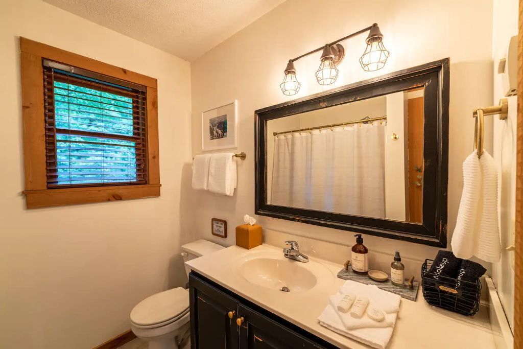 A bathroom with a large mirror, a sink, and a toilet. There is a window with blinds, a towel rack, and a small framed picture on the wall.