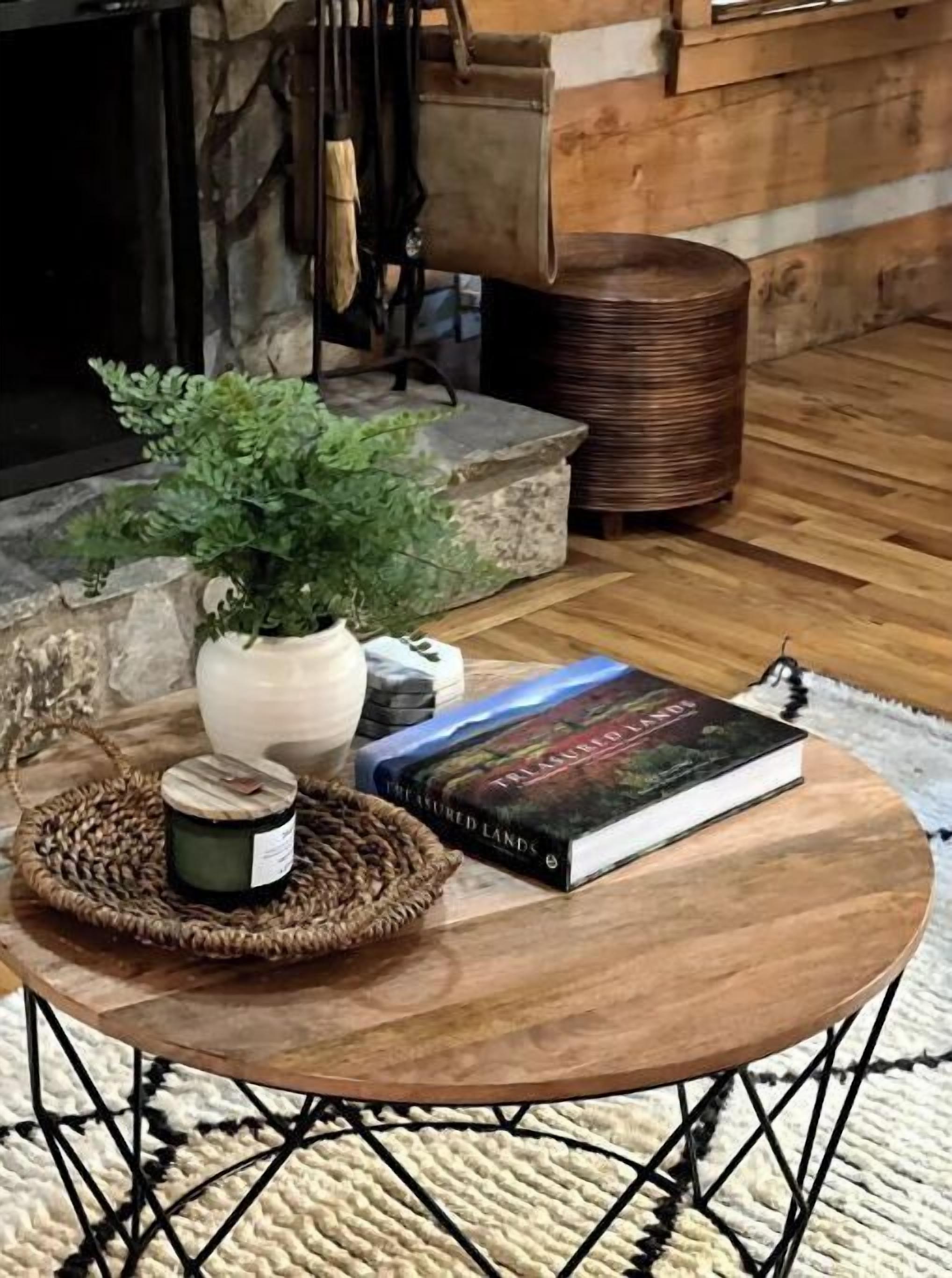 Een ronde houten tafel met een geweven onderzetter, een potplant, een kaars en een boek getiteld 'The Enchanted Lands' op een geruit tapijt.