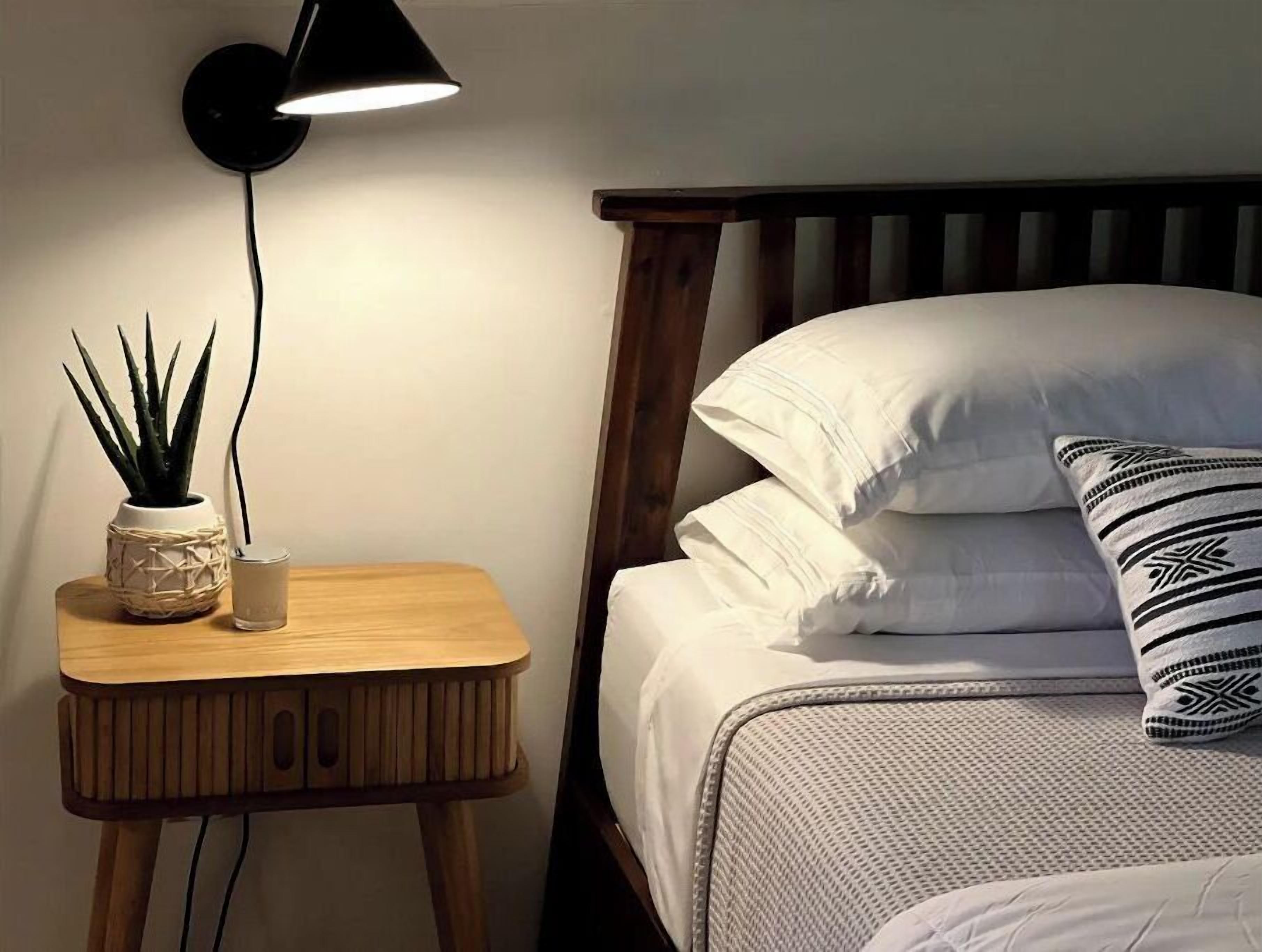 Ein ordentlich bezogenes Bett mit weißen Leinen und gemusterten Kissen, ein kleiner Nachttisch mit einem Topf und einem Glas, sowie eine Wandlampe.