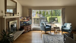 A living room with a fireplace, a television displaying a Marvel series, and a view of a garden through large windows. - Charming City Home with Professional WFH Setup & AC (Bellevue)
