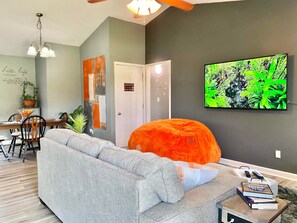 Un salon avec un canapé gris, un grand pouf orange, un coin repas avec une table et des chaises, un tableau représentant un feuillage verdoyant et une pancarte « Bienvenue » sur la porte.
