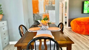A dining area with a wooden table set for four, chairs, a plant, and a painting on the wall.