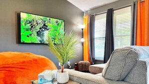 A modern living room with a grey sofa, a large orange bean bag, a wall-mounted TV, and a potted plant.