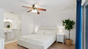 Quarto com cama, mesas de cabeceira, ventilador de teto, planta em vaso e janela com cortina azul.