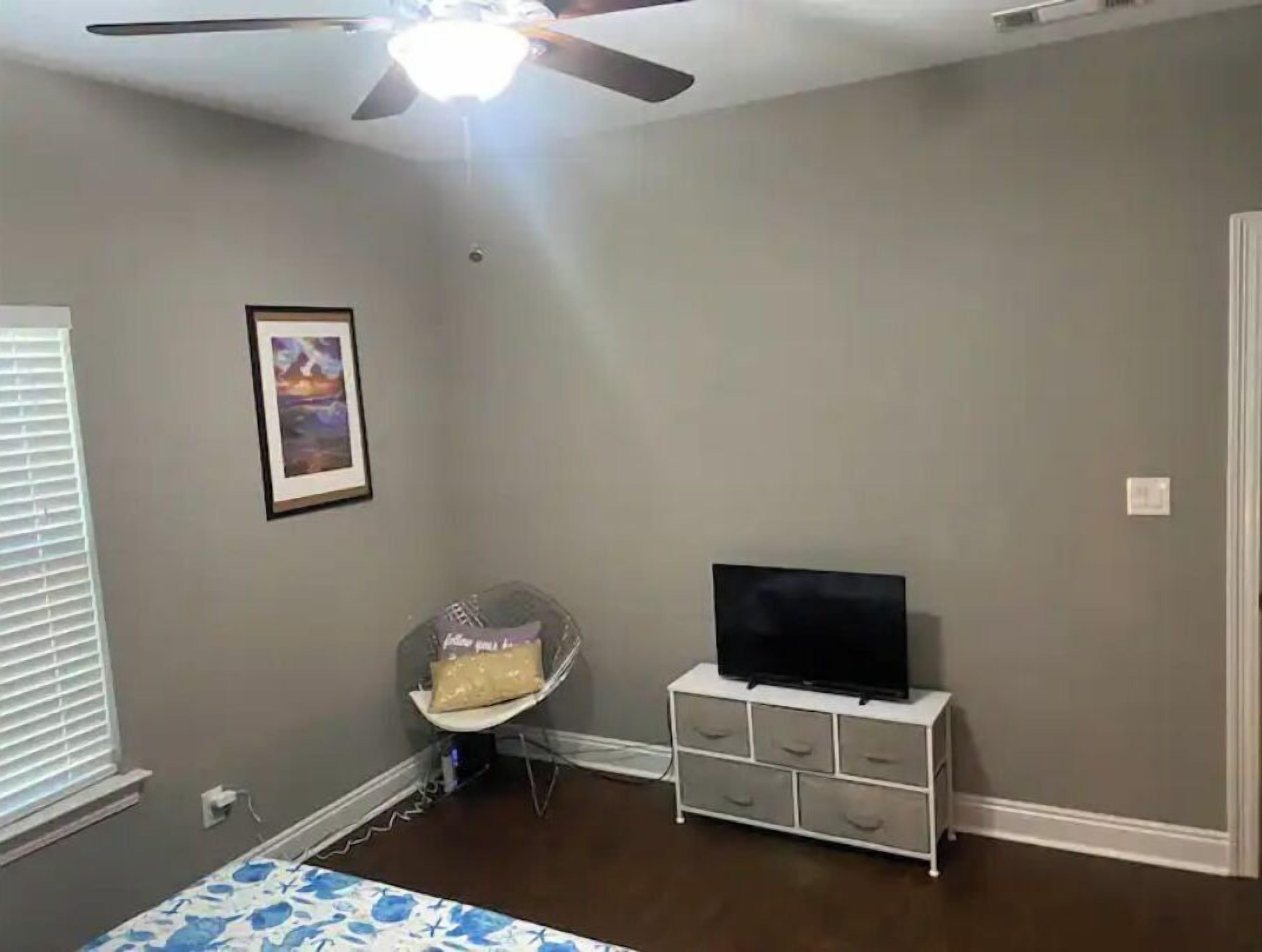 Ein Schlafzimmer mit Deckenventilator, Fernseher auf einer Kommode, einem Sessel und einem gerahmten Bild an der Wand.
