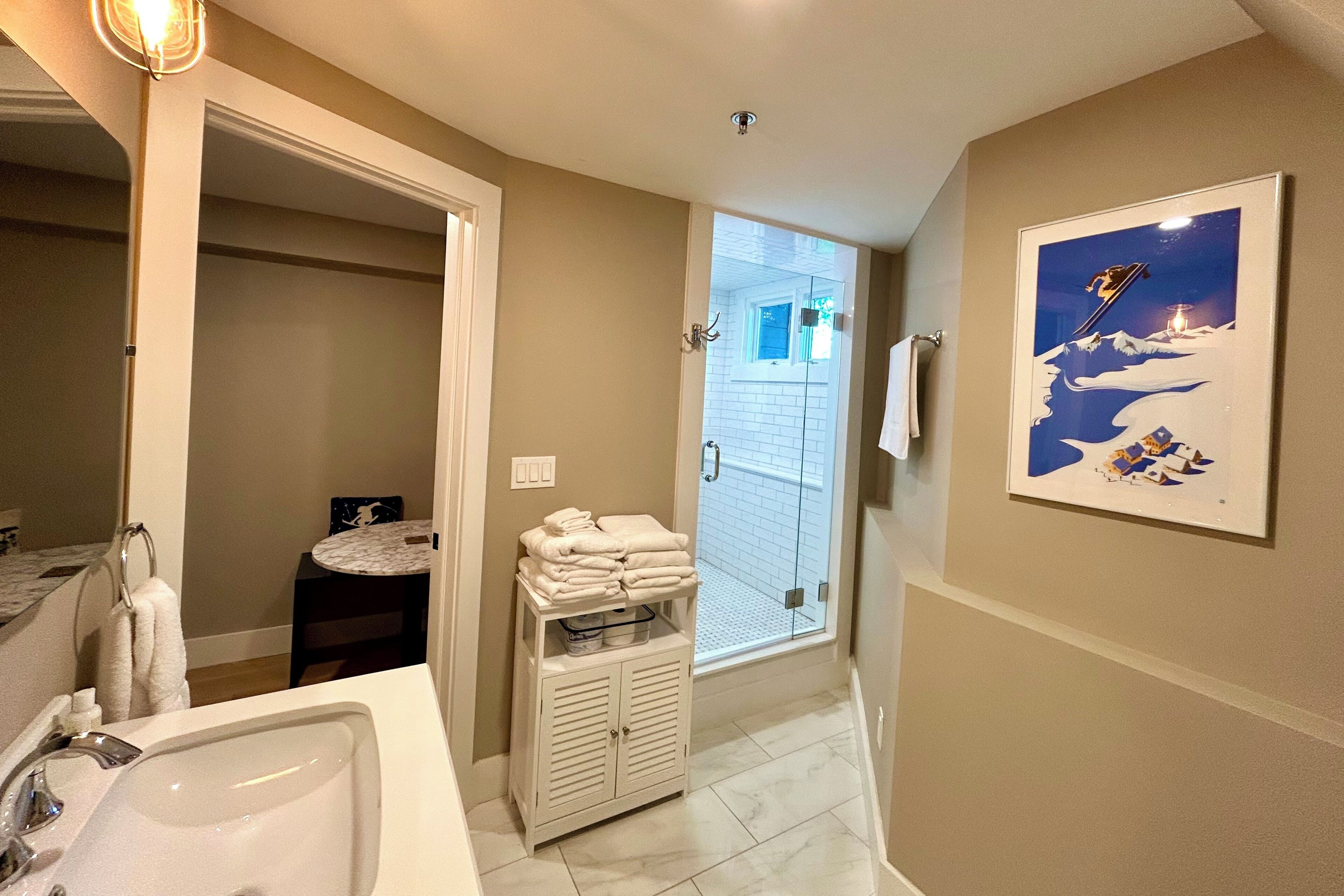 Un baño con lavamanos, espejo y toallero. Hay una puerta que da a otra habitación.