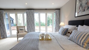 A bedroom with a large bed, a bedside table with a lamp, a chair, and a view through a glass door. - Bird Cottage, Burgh (Woodbridge)