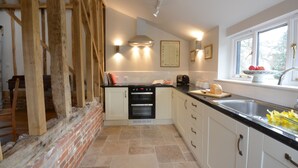 A kitchen with white cabinets, a black oven, and a window with a view of trees. - Bird Cottage, Burgh (Woodbridge)
