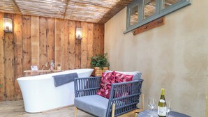 A wooden-floored room with a bathtub, a blue chair with a red pillow, and a table with a wine bottle and glass.