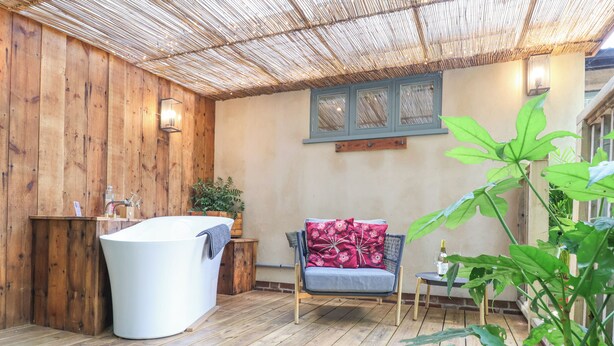 A wooden-floored bathroom with a freestanding bathtub, a wooden bench with a floral cushion, and a potted plant.