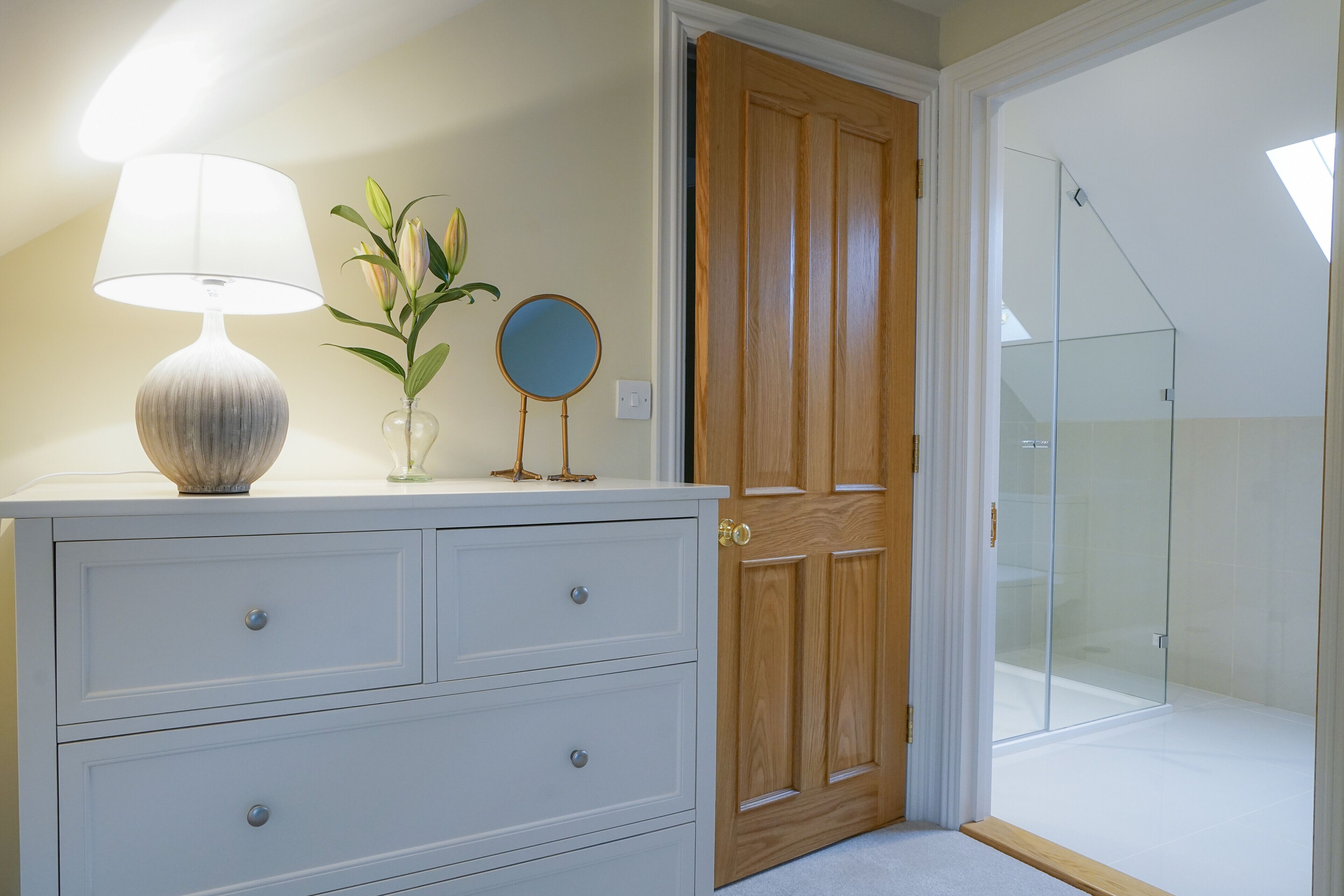 A bedroom with a wooden door, a glass shower, and a dresser with drawers and a lamp.