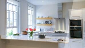 A modern kitchen with white cabinetry, a central island, a built-in oven, and a window with blinds. - 10 Church Walk, Aldeburgh (Aldeburgh)