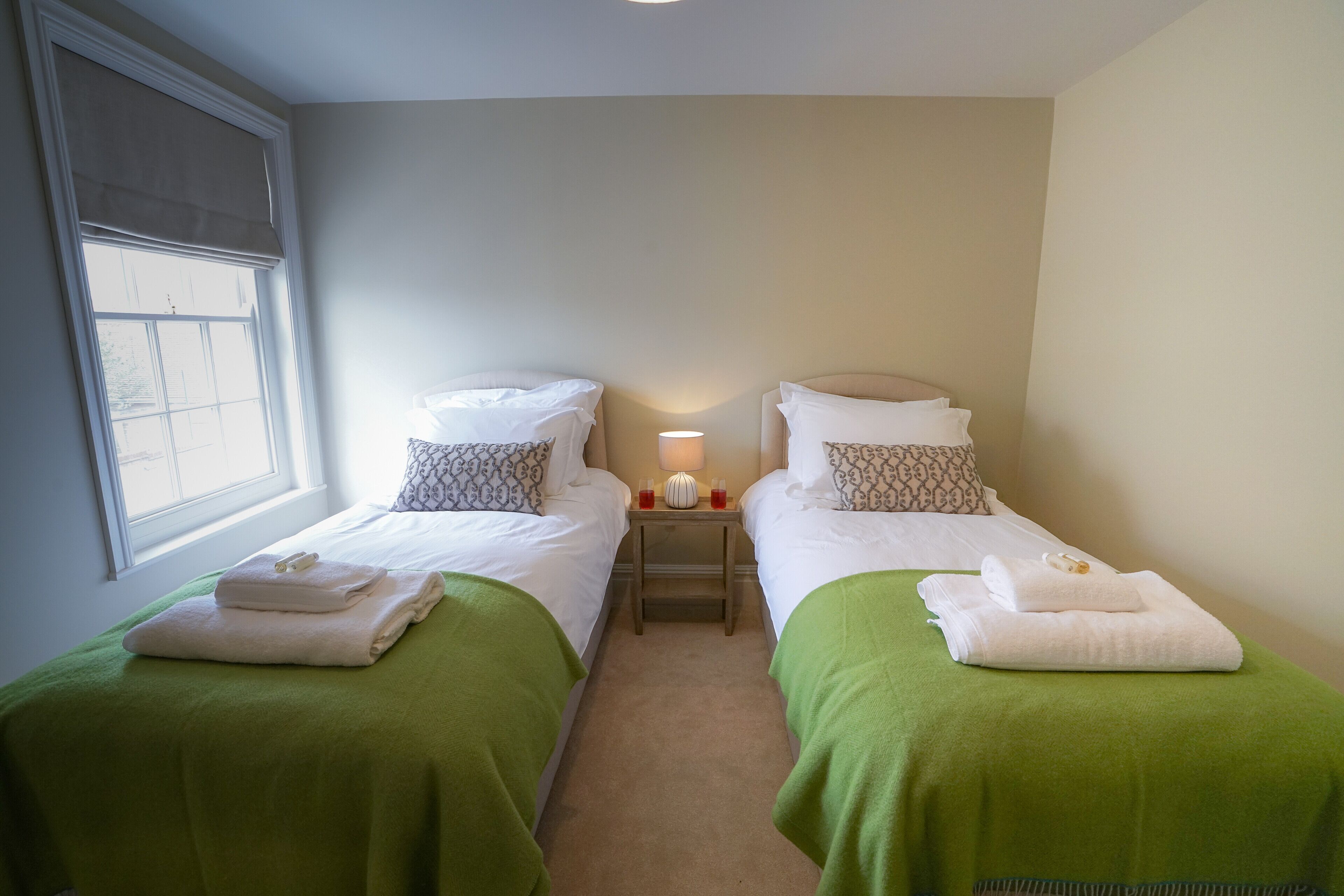 A twin room with two beds, each with green bedspreads and white linens, a bedside table with a lamp, and a window with blinds.
