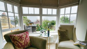 A corner room with two armchairs, a small table with a plant and wine glasses, and a view of buildings through large windows. - 10 Church Walk, Aldeburgh (Aldeburgh)