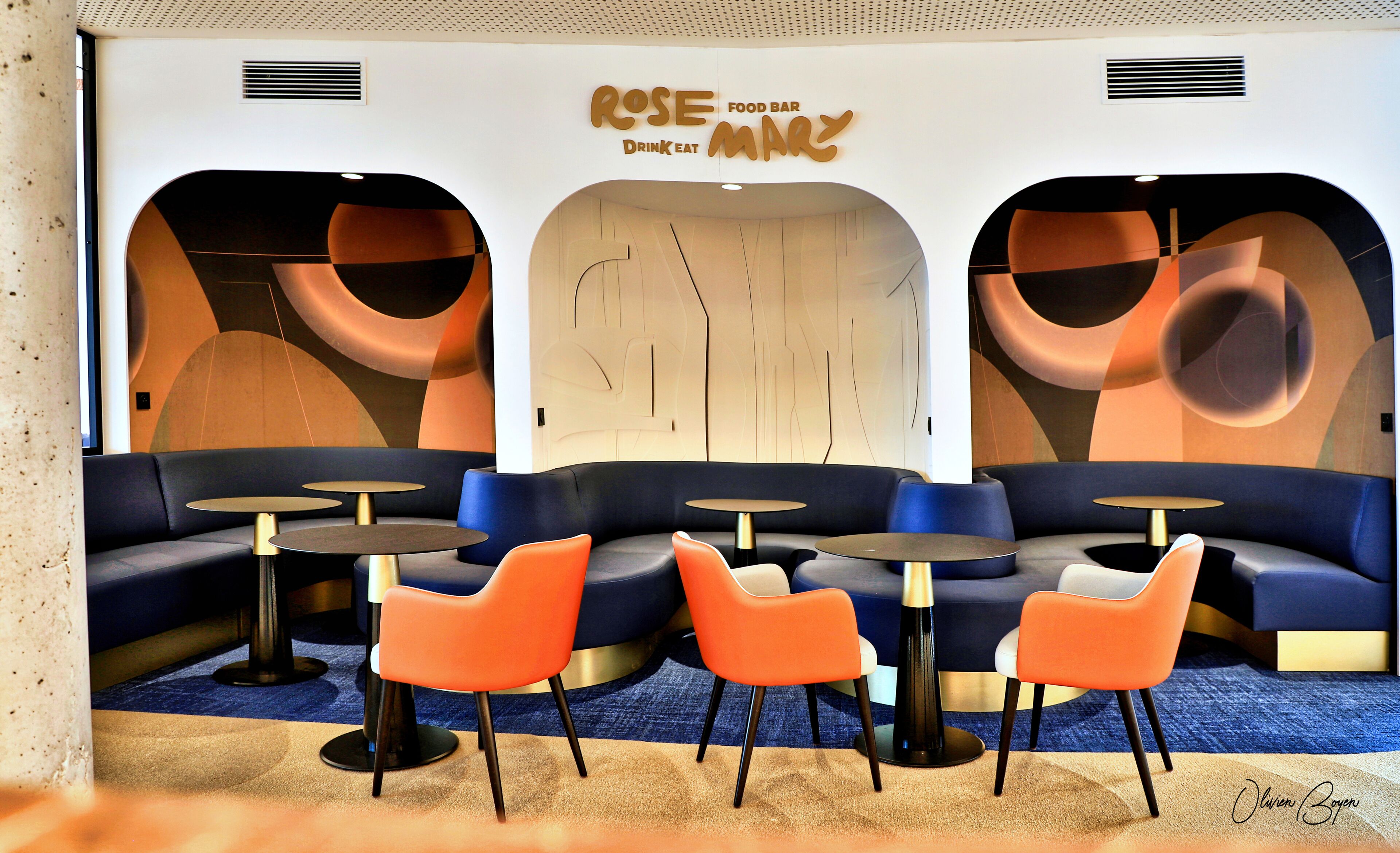 Um interior moderno com área de estar, incluindo sofás e mesas redondas, uma parede com arte abstrata e uma placa indicando “ROSE FOOD BAR”.
