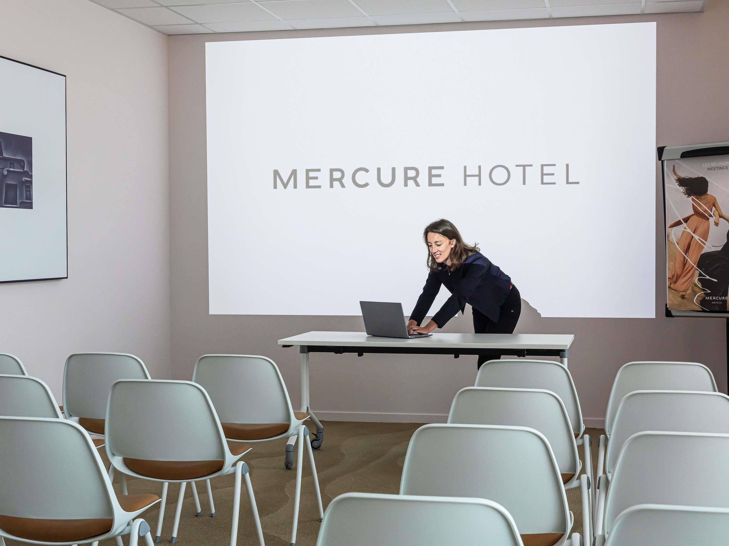 A conference room with a projection screen displaying 'MERCURE HOTEL' and a person using a laptop.