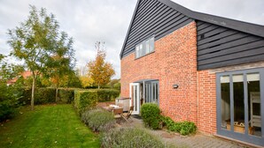 A modern house with a brick exterior, a black roof, and a glass door leading to a patio area with a table and chairs. - Three Chantry Barns, Orford (Woodbridge)