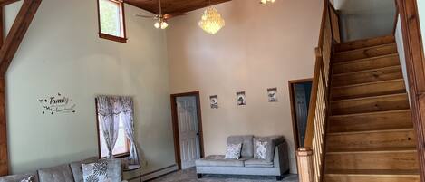 A spacious living area with a high ceiling, wooden beams, a staircase, a sofa set, a coffee table, and a chandelier.