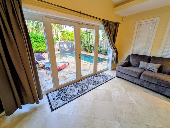 A living room with a grey sofa, a large sliding glass door leading to a pool area, and a patterned rug on the floor.