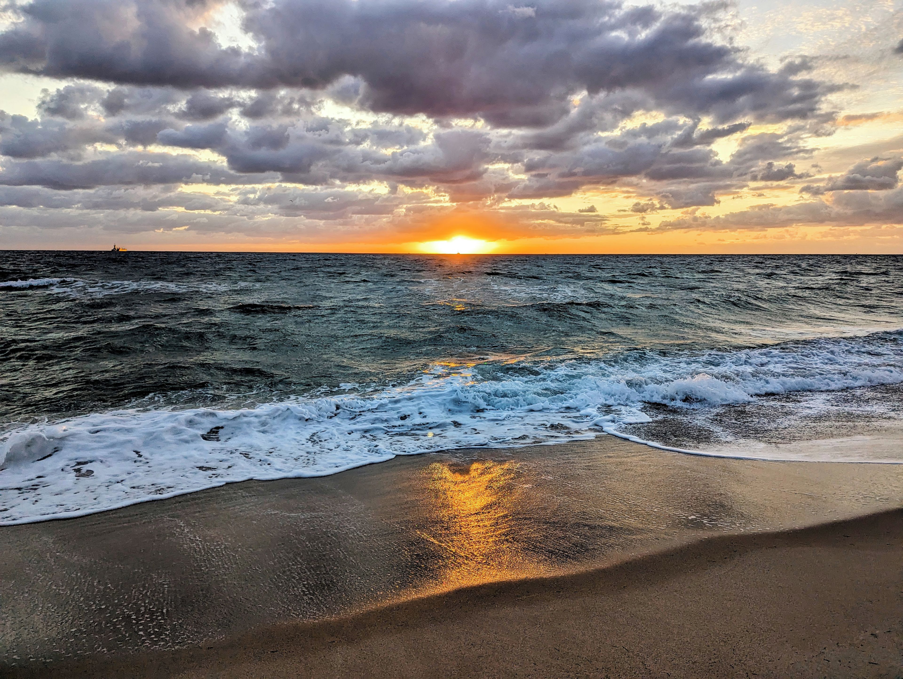 A beach with waves approaching the shore, a sunset in the background, and a distant boat.