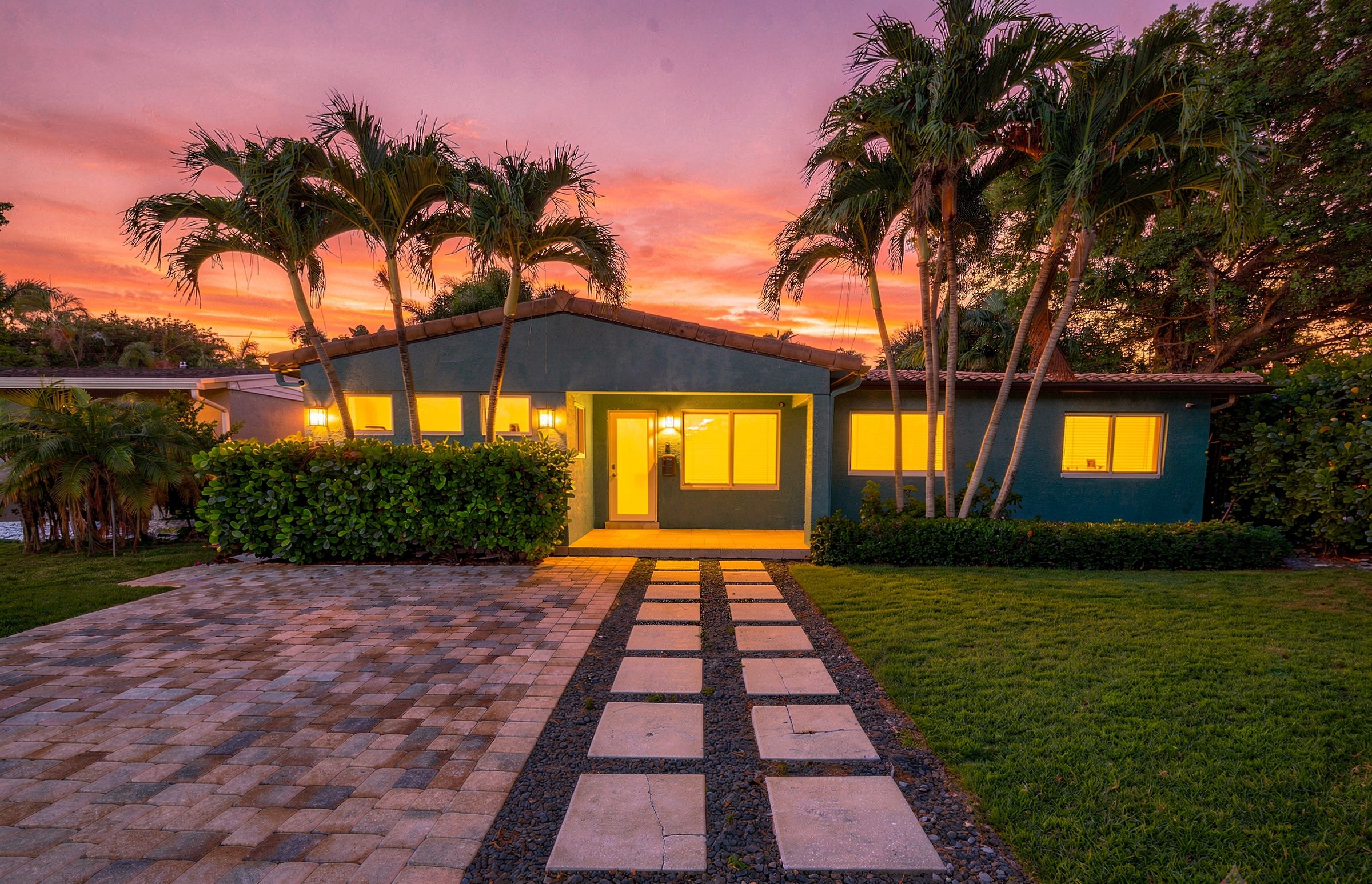 A modern house with a paved driveway leading to a front door, surrounded by palm trees and a well-maintained lawn.