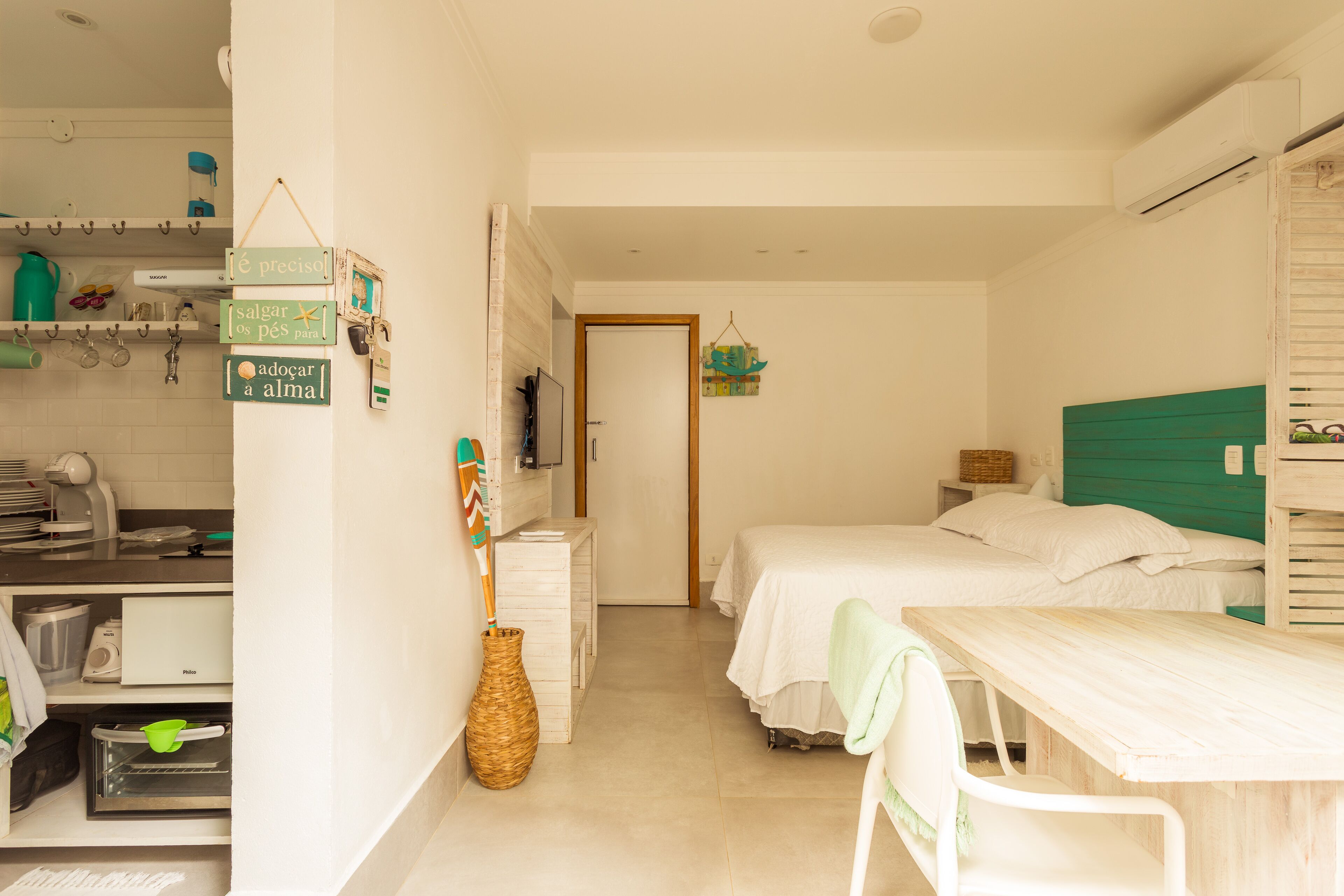 Habitación de hotel compacta con kitchenette, una cama con cabecera verde, una mesita y una silla.