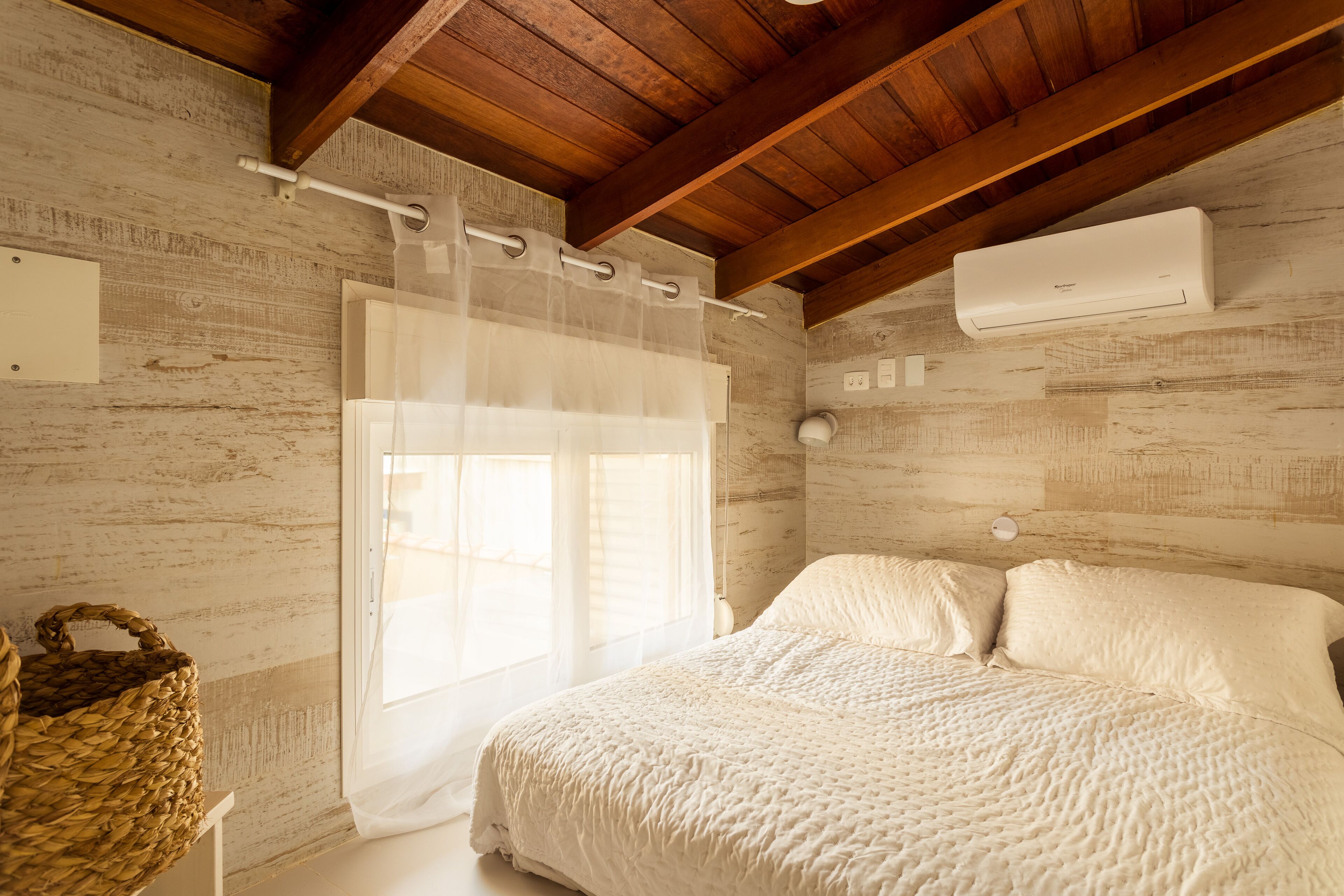 A bedroom with a wooden ceiling, a bed with white bedding, a window with a sheer curtain, and an air conditioning unit on the wall.