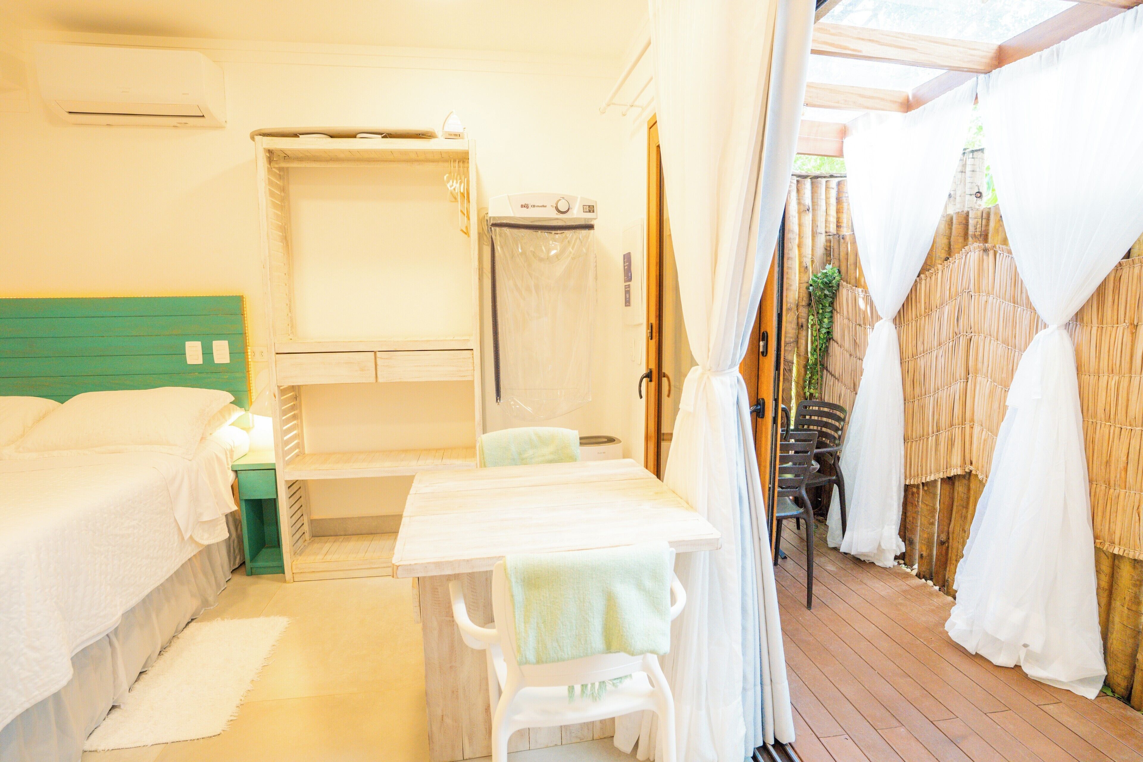 A room with a bed, a table with a chair, a shower area, and a view of the outdoors.