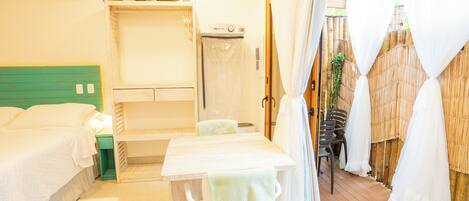 A room with a bed, a table with a chair, a shower area, and a view of the outdoors.