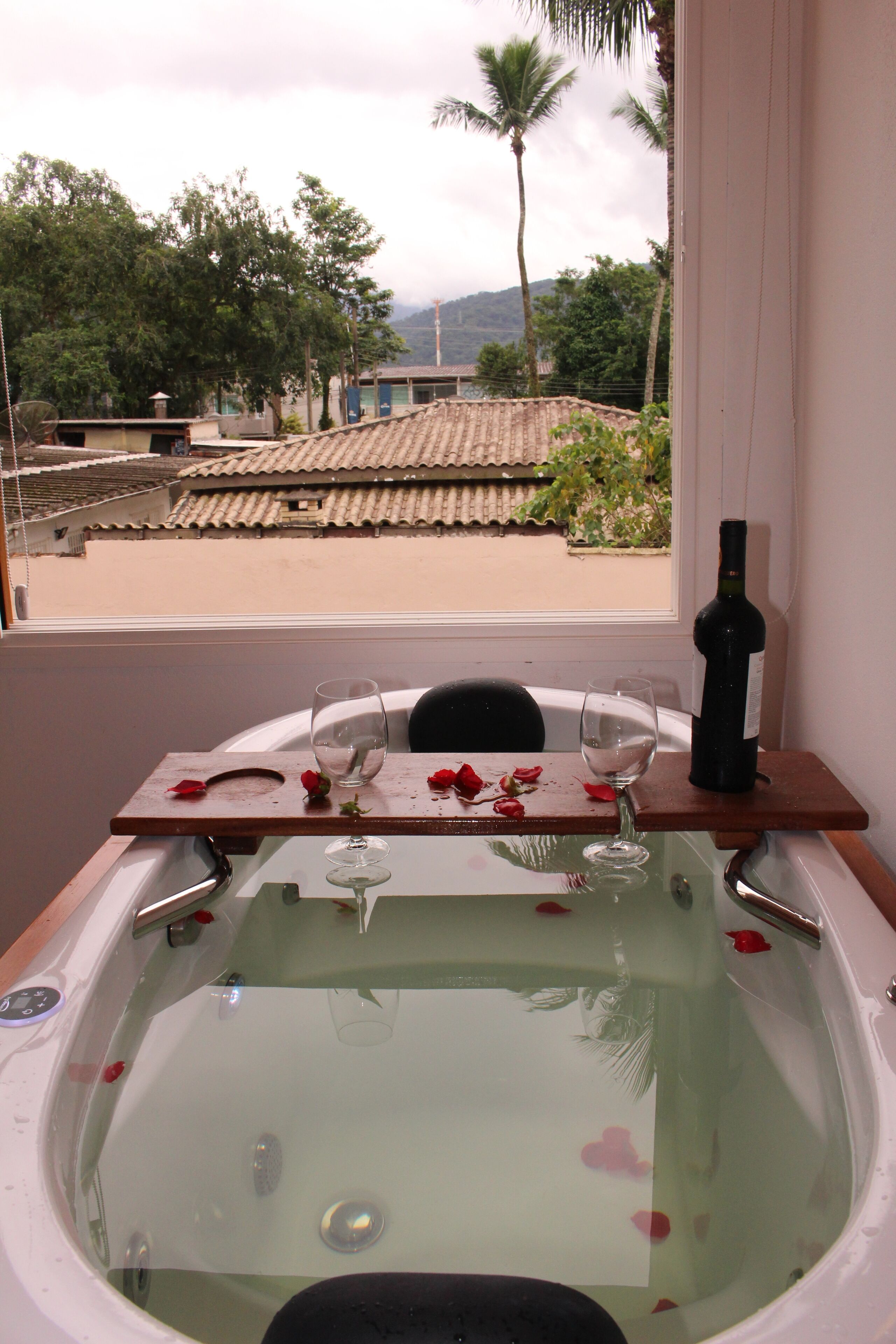 A bathtub with rose petals and a wine bottle, a wooden tray, and a glass.