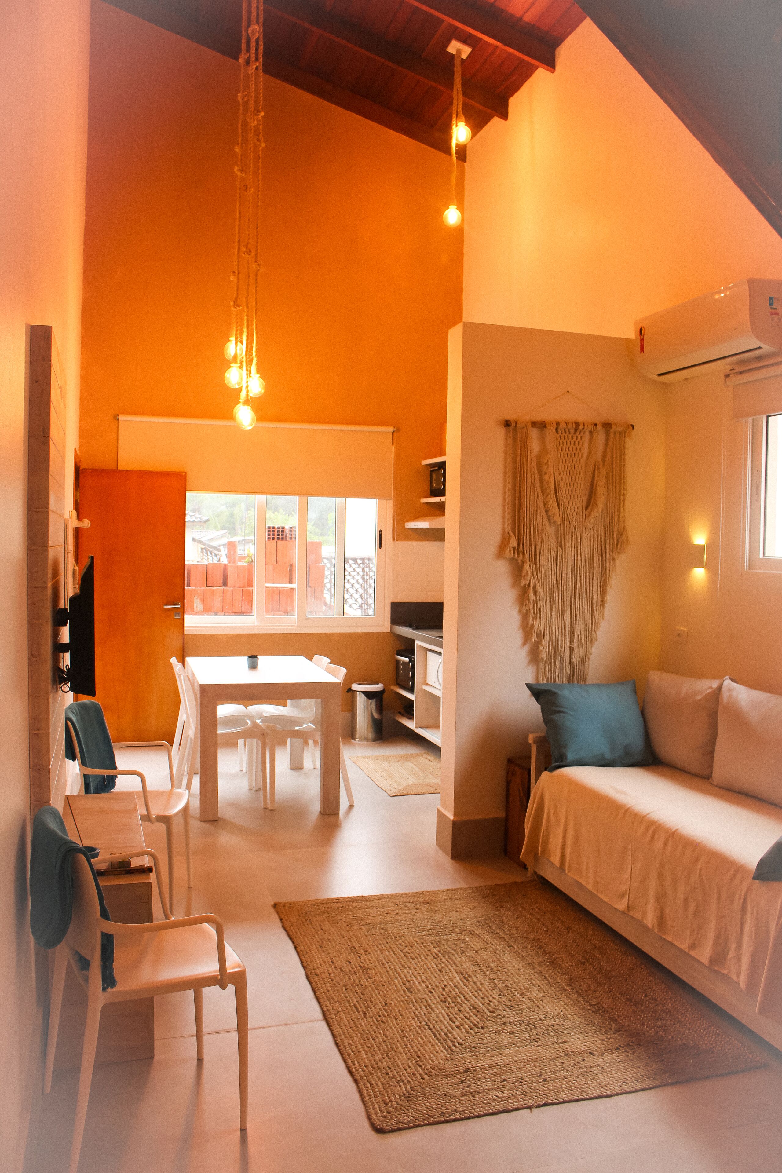 Ein gemütliches Wohnzimmer mit einer Couch, einem Esstisch und Stühlen. Im Hintergrund ist eine Küchenzeile mit einem Fenster und einer Wanddekoration aus Makramee zu sehen.