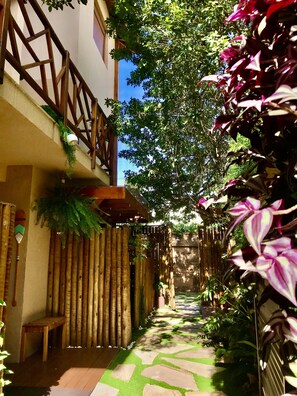 Un étroit sentier bordé de clôtures en bambou et d’une végétation luxuriante, qui mène à un bâtiment doté d’un balcon en bois.