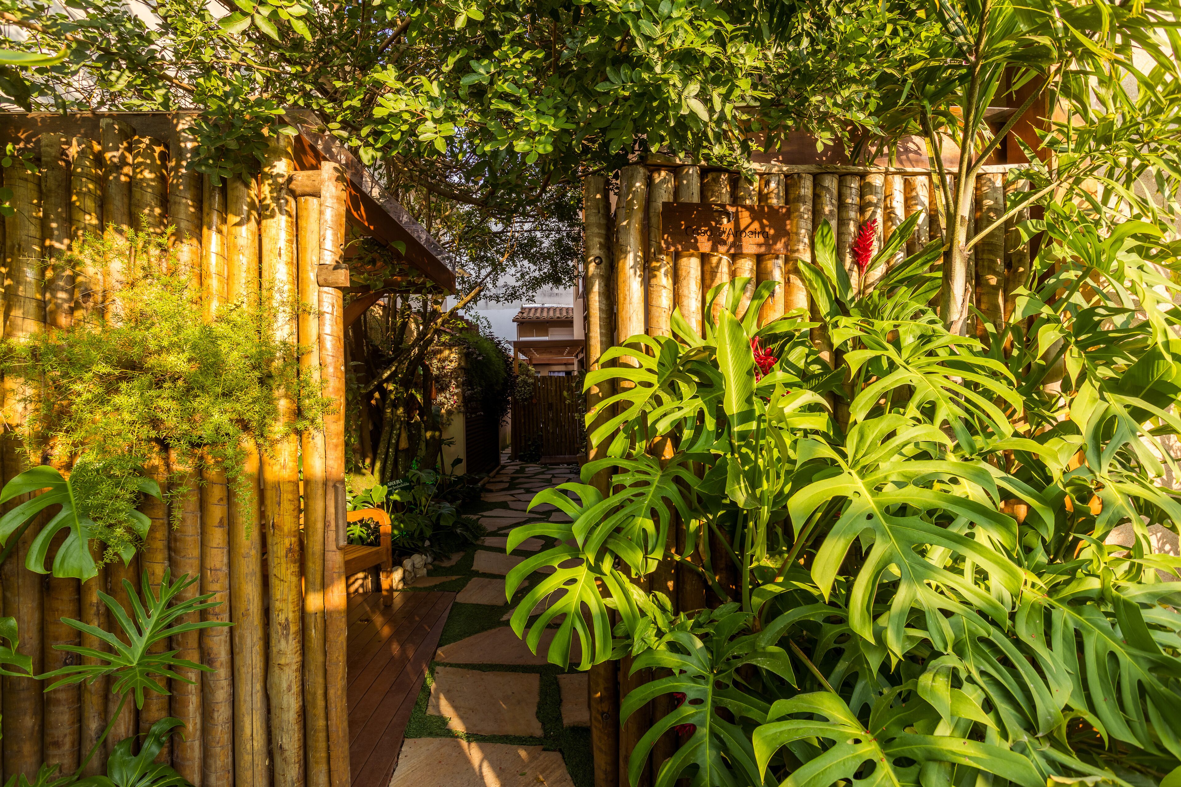 Un sentier de jardin qui mène à une maison traditionnelle, entourée d’une clôture en bambou et d’une végétation luxuriante.