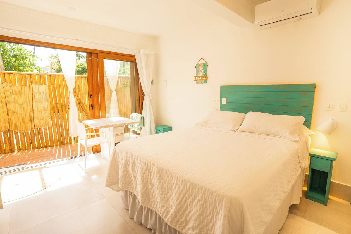 Une chambre avec un grand lit, un bureau et une chaise, une porte-fenêtre coulissante donnant sur un balcon, et une petite table de chevet verte.